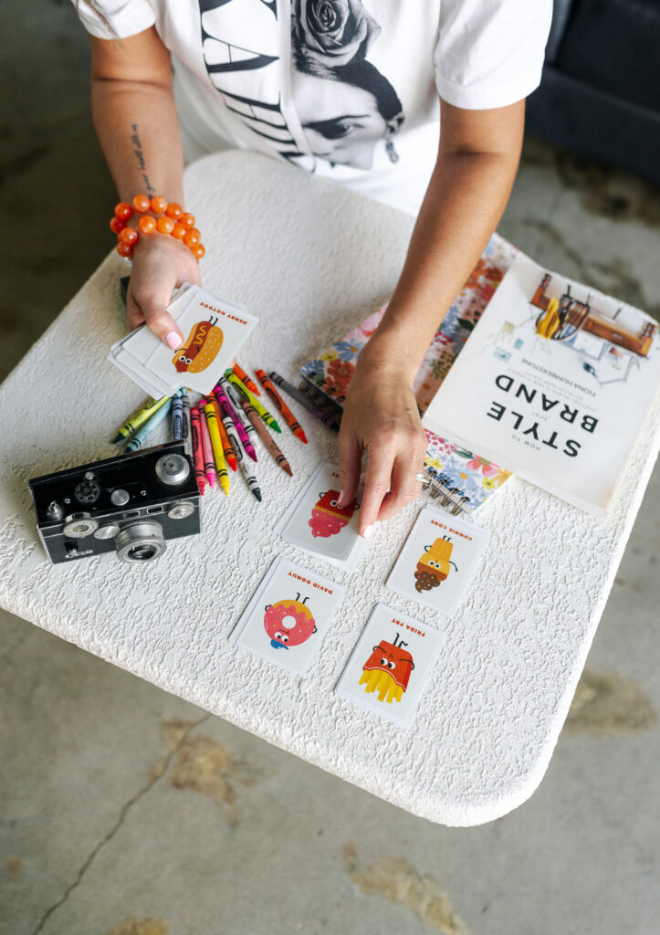 Close-up of Desiree’s hands at a coffee table, playing a kids’ card game surrounded by crayons, a vintage film camera, and a branding visuals book, blending family life with creative entrepreneurship.